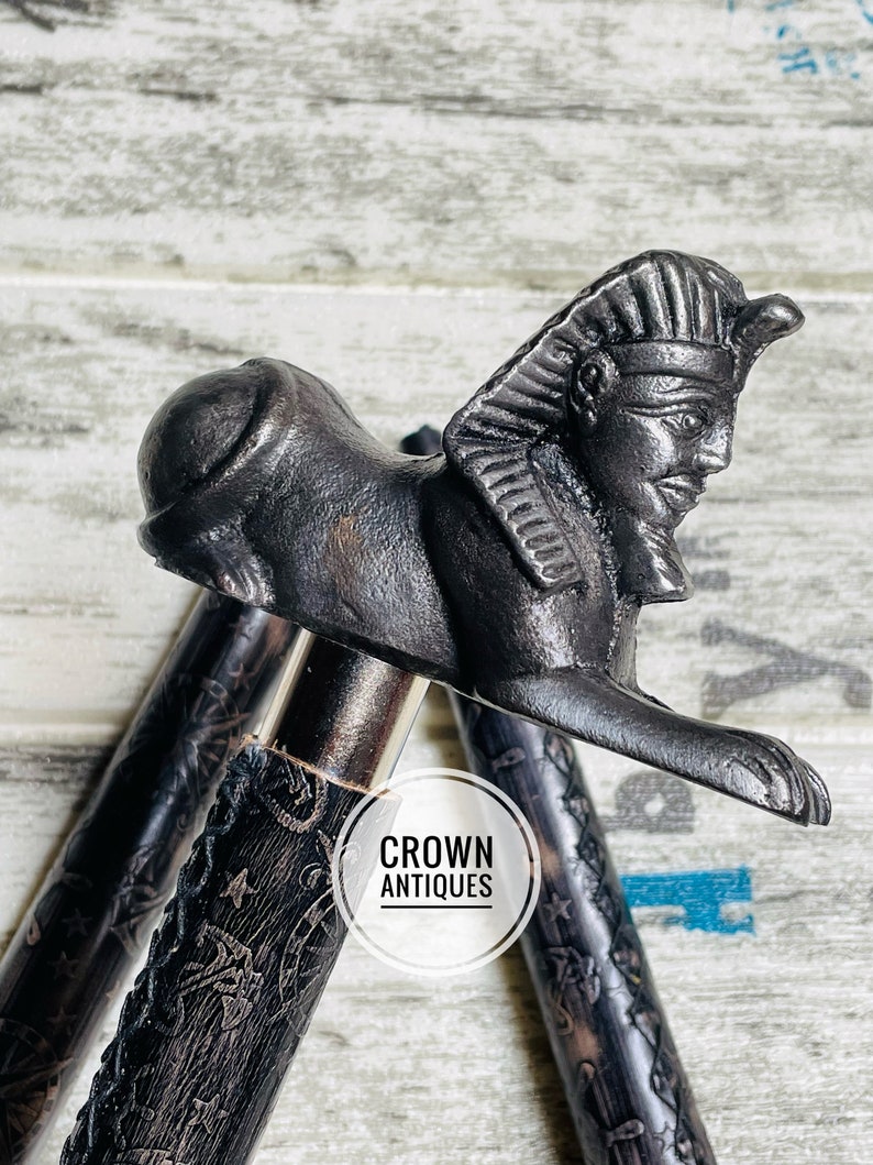 May include: A black walking stick with a detailed carved design and a decorative metal head shaped like a sphinx. The head is detailed with a pharaoh's headdress and facial features. The text "CROWN ANTIQUES" is visible on the stick.