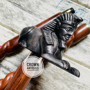 May include: A wooden walking stick with a black metal Egyptian sphinx head handle. The handle is detailed with a headdress and facial features. The stick is made of dark wood with a natural grain pattern. The text "CROWN ANTIQUES" is visible on a white circle sticker on the stick.