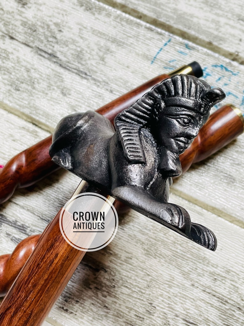 May include: A wooden walking stick with a black metal Egyptian sphinx head handle. The handle is detailed with a headdress and facial features. The stick is made of dark wood with a natural grain pattern. The text "CROWN ANTIQUES" is visible on a white circle sticker on the stick.