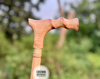 May include: A light brown wooden walking cane with a curved handle. The handle has a carved design for grip and comfort. The cane has a decorative gold band near the top. The logo "CROWN ANTIQUES" is visible on the cane.