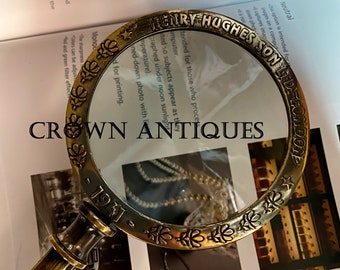 May include: A brass magnifying glass with a decorative floral design around the rim. The glass is dated "1941" and has the inscription "Henry Hughes Son Ltd London" around the top. The magnifying glass is on a white surface with a book in the background.