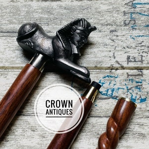 May include: Two wooden walking sticks with decorative handles. One stick has a twisted design and the other has a handle shaped like a sphinx. The sticks are on a white wooden surface. The text "CROWN ANTIQUES" is visible in the image.