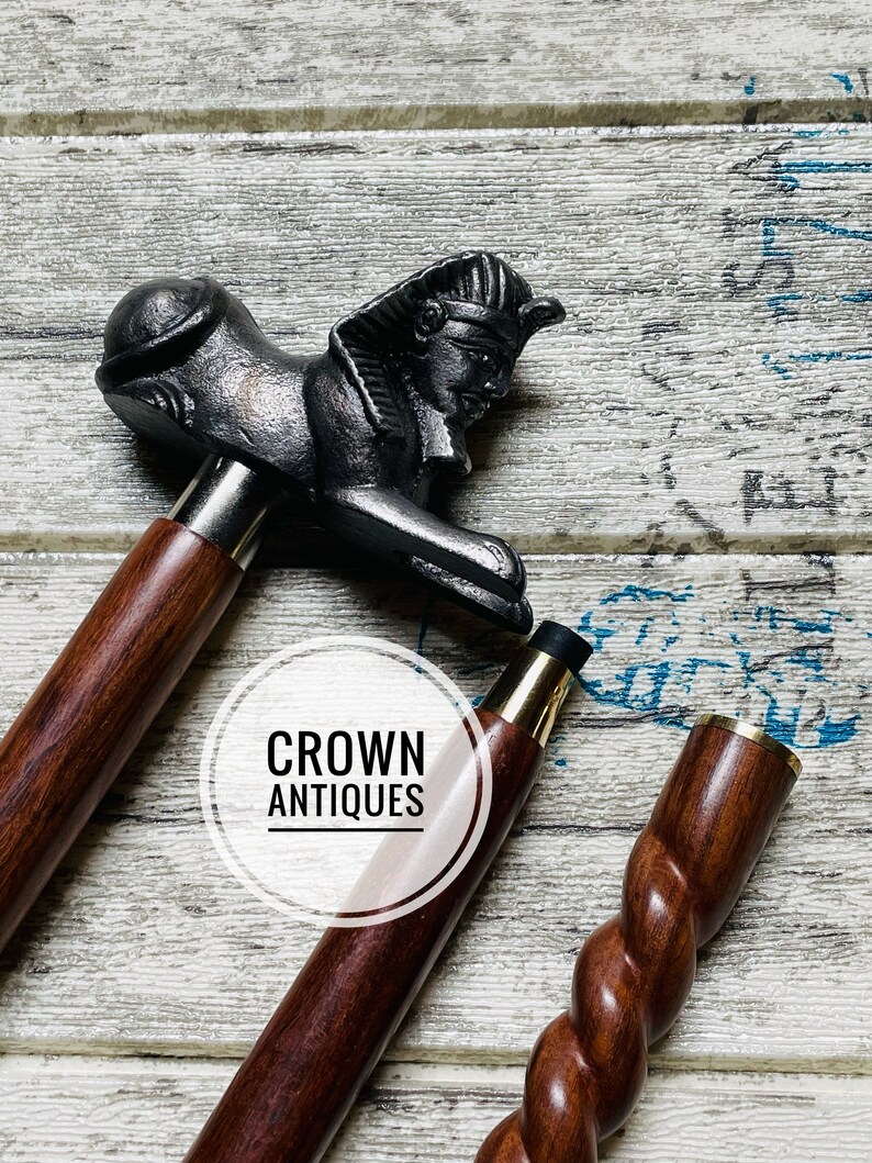 May include: Two wooden walking sticks with decorative handles. One stick has a twisted design and the other has a handle shaped like a sphinx. The sticks are on a white wooden surface. The text "CROWN ANTIQUES" is visible in the image.