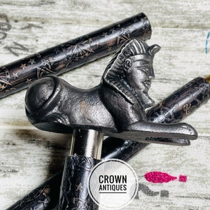 May include: A black walking stick with a detailed design and a decorative metal handle shaped like an Egyptian sphinx. The handle is a dark gray color and has a detailed face and body. The stick is made of wood and has a black finish. The text "CROWN ANTIQUES" is visible on the stick.