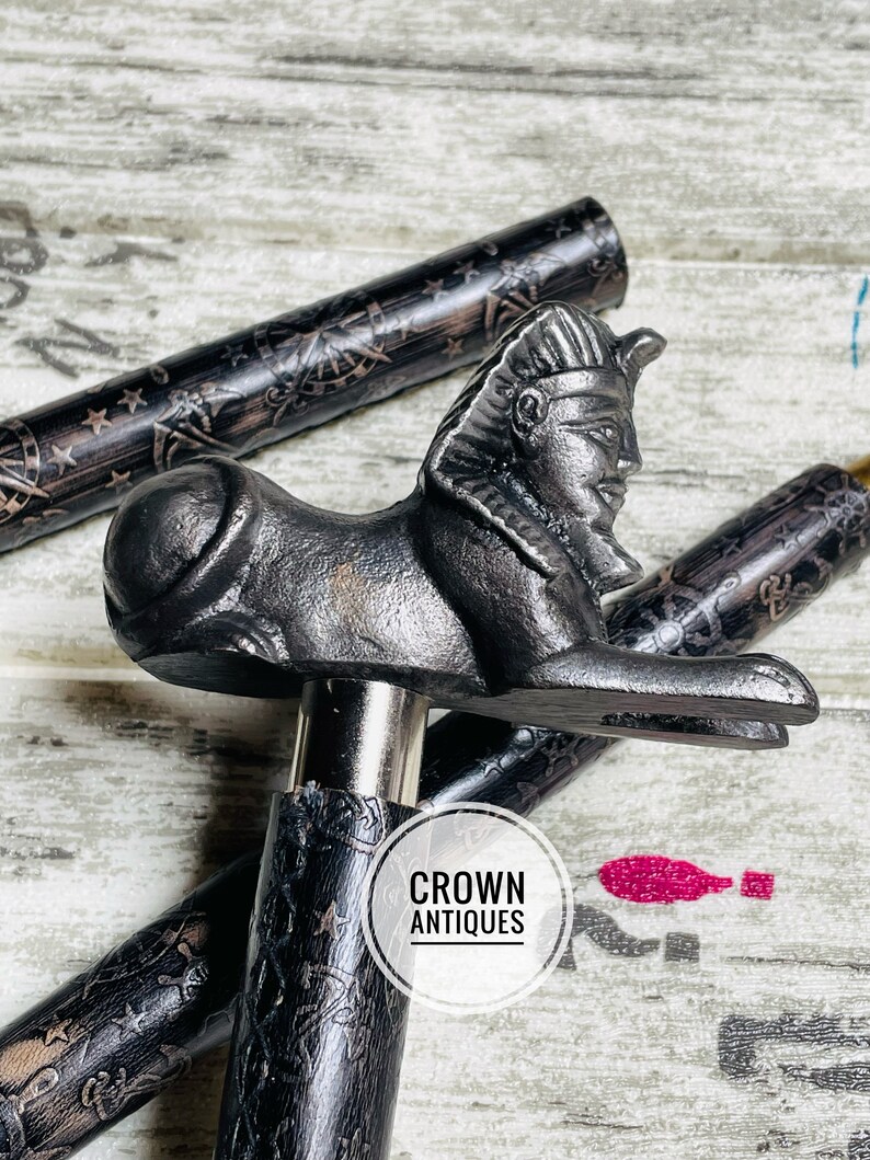 May include: A black walking stick with a detailed design and a decorative metal handle shaped like an Egyptian sphinx. The handle is a dark gray color and has a detailed face and body. The stick is made of wood and has a black finish. The text "CROWN ANTIQUES" is visible on the stick.