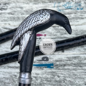 May include: A black and silver walking stick with a crow head handle. The handle is detailed with silver feathers and the stick has a black and silver patterned shaft.