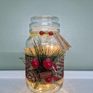 May include: A clear glass jar with a lid, decorated with a festive arrangement. The jar is adorned with red berries, pine needles, a cinnamon stick, and a plaid ribbon. A tag that says "HANDMADE WITH LOVE" hangs from the jar.