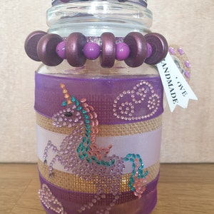 May include: A decorative glass jar with a lid, adorned with purple ribbons, burlap, and a rhinestone unicorn design. The jar is embellished with wooden beads and a tag that reads "HANDMADE LOVE".