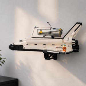 Può includere: Modello bianco e nero di uno space shuttle montato su una parete. Il modello ha un serbatoio esterno argentato e una piccola figura di astronauta. Lo shuttle è esposto con le ali spiegate. È un oggetto da collezione.