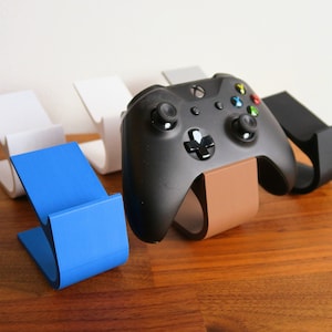 May include: Four different coloured 3D printed stands for a video game controller. The stands are white, grey, blue, and brown. A black video game controller is sitting on the brown stand.