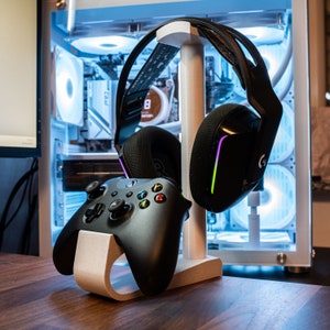 Pode incluir: Um suporte branco com um controlador de videogame preto e cinza e um fone de ouvido para jogos preto com detalhes roxos e verdes. O suporte está em uma superfície de madeira.