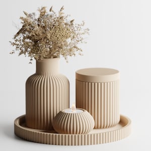 May include: A beige ribbed set consisting of a vase filled with dried flowers, a lidded canister, and a small tea light holder, all arranged on a matching circular ribbed tray. The items feature a consistent vertical fluted texture.