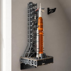 Può includere: Modello dettagliato di un razzo e di una torre di lancio, prevalentemente grigi, arancioni e bianchi, montato a parete. Il razzo presenta booster arancioni e sezioni bianche, con la torre di lancio costruita con componenti grigi. Replica di un sistema di lancio spaziale.