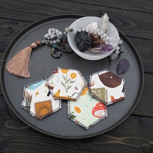 May include: A collection of five hexagonal fabric coasters with various nature-themed designs, including a sun, flowers, and mushrooms. The coasters are arranged on a dark gray tray, alongside a bowl of crystals and a beaded necklace.