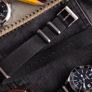 May include: A black nylon watch strap with a silver buckle. The strap is laid on a dark denim fabric background.