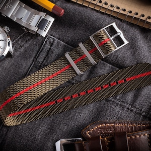 Op de afbeelding: Een groen en rood gestreept nylon horlogebandje met een zilveren gesp. Het bandje ligt op een donkergrijs stofoppervlak.