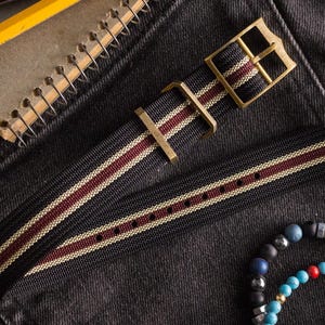 May include: A black watch strap with burgundy and cream stripes and brass hardware. The strap is made of a woven material and has a classic design. The watch strap is shown on a dark denim background.