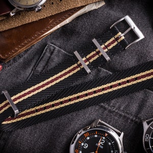 May include: Black watch strap with white and burgundy stripes. The strap is made of nylon and has a silver buckle.