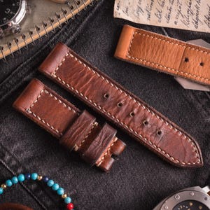 May include: Close-up of brown leather watch straps with white stitching. The straps are made of distressed leather and have multiple holes for adjustment. The background includes a watch, a notebook, and a bracelet.