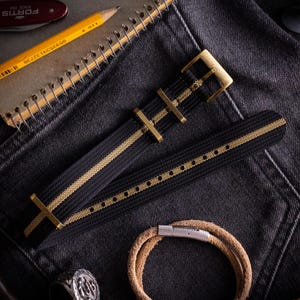 May include: A black nylon watch strap featuring a central tan stripe and brass-toned metal hardware, including a buckle and two keepers. The strap is woven with a textured finish and has a series of punched holes for adjustment along the length.