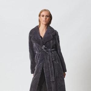 May include: A long, grey fur coat with a shawl collar and a self-tie belt. The coat is worn by a model with long blonde hair and is styled with black knee-high boots.