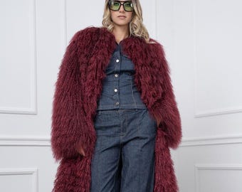 Puede incluir: Una mujer con un abrigo de piel sintética burdeos sobre un mono vaquero y pantalones de pernera ancha. El abrigo es largo y peludo. El conjunto vaquero tiene una parte superior abotonada y pantalones de cintura alta. Lleva gafas de sol oscuras.