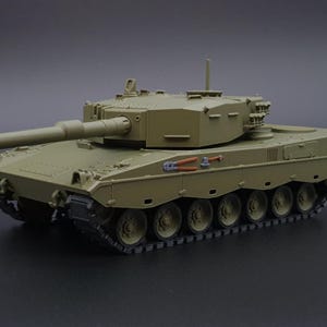 Könnte beinhalten: Ein maßstabsgetreues Modell eines Kampfpanzers in mattem Olivgrün. Der Panzer verfügt über ein langes Hauptgeschützrohr, einen drehbaren Turm und detaillierte Ketten mit mehreren Laufrollen. Kleine orangefarbene und silberne Werkzeuge sind an der Seite angebracht.