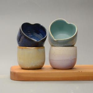 May include: Four small ceramic cups in shades of blue, yellow, and lavender, stacked on a wooden tray. The top cups have heart-shaped openings. The cups are handmade and have a rustic, artisanal appearance.
