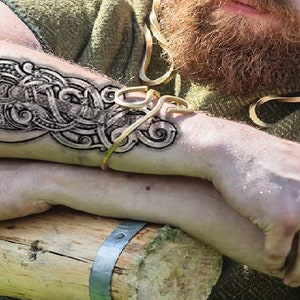 May include: A man's arm with a detailed black and white tattoo of a mythical creature with intricate knotwork. The tattoo is on the forearm and extends to the wrist. The man is wearing a green shirt and a gold bracelet.
