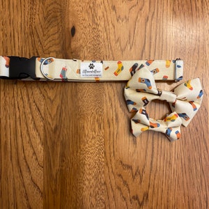 May include: A dog collar and bow tie set with a cream-colored background and a repeating pattern of colorful socks. The collar has a black plastic buckle and a metal D-ring. The bow tie is pre-tied.