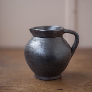Puede incluir: Una pequeña jarra de cerámica negra con cuerpo redondeado y asa. La jarra está sobre una superficie de madera.