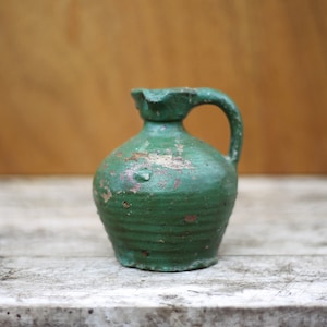 Peut inclure: Une petite cruche en céramique verte avec un corps rond et une anse. La cruche a un aspect usé et rustique avec des imperfections visibles dans le glaçage.