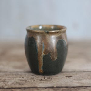 Op de afbeelding: Een kleine, handgemaakte keramische vaas met een afgeronde body en een licht uitlopende rand. De vaas heeft een donkergroene basis met bruin en tan glazuur dat langs de zijkanten druipt. De binnenkant is lichtbruin. De vaas staat op een houten oppervlak.