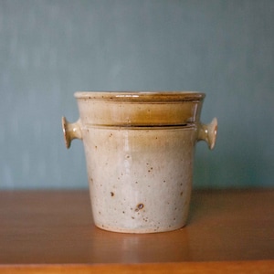 Może przedstawiać: Beige pojemnik ceramiczny z dwoma małymi uchwytami po bokach. Pojemnik ma pstrokatą glazurę i brązową krawędź.