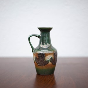 May include: A green ceramic vase with a brown and black design. The vase has a rounded body and a short neck with a handle.