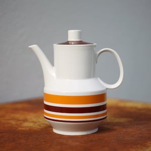 Peut inclure: Une cafetière en céramique blanche avec un couvercle brun et des rayures orange et brunes autour du bas.