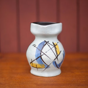 Peut inclure: Petit vase en céramique blanche avec intérieur noir et forme originale. Le vase est décoré de lignes abstraites bleues, jaunes et noires. Le vase est posé sur une surface en bois.