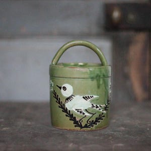 Può includere: Vaso in ceramica verde con manico, con un disegno di uccello dipinto a mano. L'uccello è bianco con dettagli neri, su uno sfondo di fogliame verde. Il vaso è cilindrico e ha un manico curvo.
