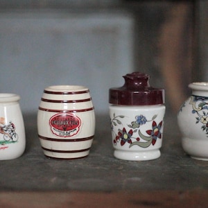 Op de afbeelding: Vier kleine, decoratieve keramische potten op een rij. De potten zijn wit met verschillende ontwerpen, waaronder een paardenwagen, een vatvorm met tekst, bloempatronen en een decoratief bloemmotief. De potten staan op een donkere ondergrond.