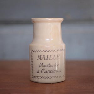 May include: Cream-colored ceramic jar with the text "MAILLE Moutarde à l'ancienne" in black script. The jar has a cylindrical shape with a slightly flared top and a decorative border around the label. The jar is on a wooden surface.