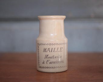 May include: Cream-coloured ceramic jar with the text "MAILLE Moutarde à l'ancienne" in black script. The jar has a cylindrical shape with a slightly flared top and a decorative border around the label. The jar is on a wooden surface.