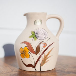 Könnte beinhalten: Eine beige Keramikkanne mit Blumenmuster. Die Kanne hat einen braunen Henkel und ist mit weißen, gelben, orangefarbenen und grünen Blumen verziert.