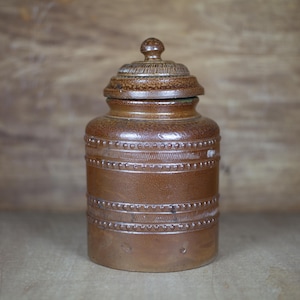 Puede incluir: Un tarro de cerámica marrón con tapa, que presenta un diseño decorativo de puntos y líneas en relieve. El tarro tiene un aspecto rústico y vintage.