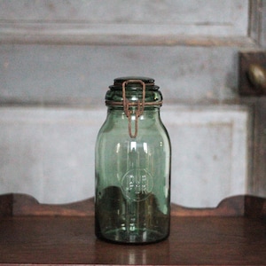 Op de afbeelding: Een groene glazen pot met een metalen sluiting. De pot heeft een afgeronde vorm en een donkere metalen deksel. De pot staat op een houten oppervlak. De pot heeft een vintage uitstraling.