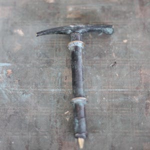 Puede incluir: Un martillo de metal antiguo con un aspecto envejecido. El martillo tiene una cabeza en forma de T y un mango cilíndrico. El metal muestra signos de desgaste y oxidación, con tonos oscuros y verdosos.