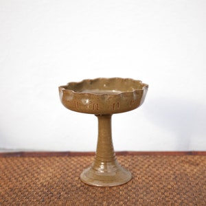 May include: A brown ceramic pedestal bowl with a scalloped edge. The bowl is sitting on a woven mat.