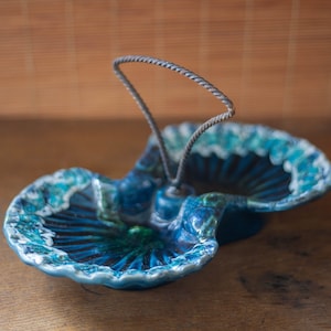May include: A blue and green ceramic serving dish with a metal handle. The dish is shaped like two seashells joined together.