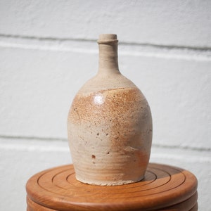 Könnte beinhalten: Eine hellbraune Keramikflasche mit rundem Körper und schmalem Hals. Die Flasche hat ein rustikales, handgemachtes Aussehen mit einer gesprenkelten Glasur.