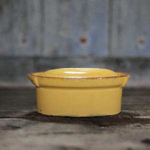 Peut inclure: Un petit plat rond en céramique jaune avec un couvercle. Le plat a un aspect légèrement vieilli avec un bord marron. L'arrière-plan est une surface en bois vieilli et un mur bleu délavé.