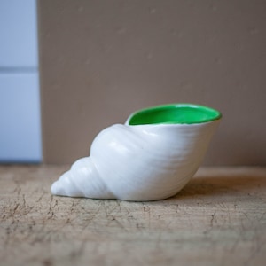 Può includere: Un vaso da fiori in ceramica bianca a forma di conchiglia con interno smaltato verde. Il vaso è appoggiato su una superficie di legno.